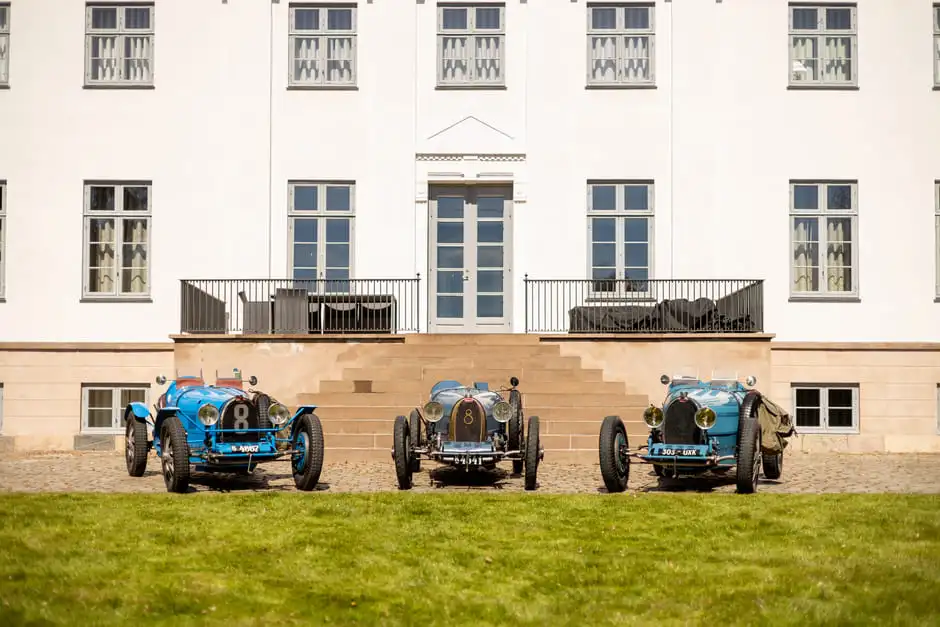 Three blue Bugatti Grand Prix cars lined up before the white manor house at Aggersvold Estate.