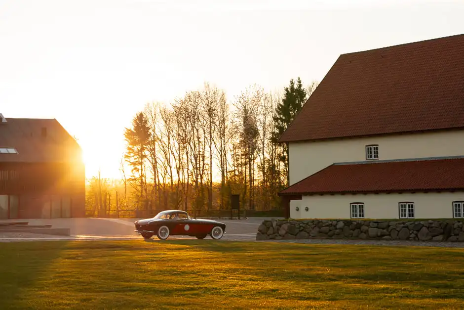 A classic red coupe crossing the grounds of Aggersvold Estate at sunset, the estate barns behind.