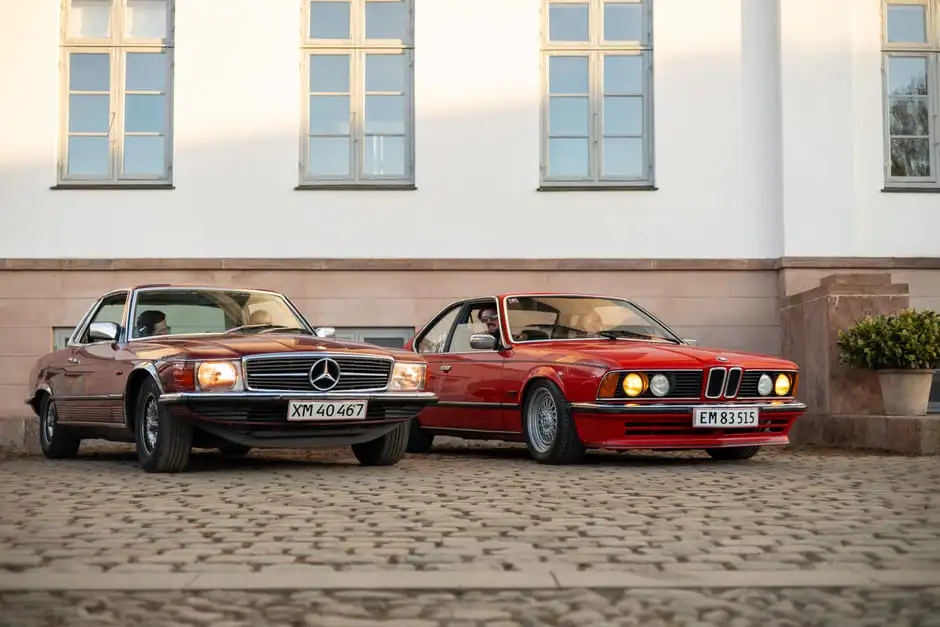 A vintage Mercedes-Benz coupe and a red BMW parked on the cobbles in front of the Aggersvold manor.