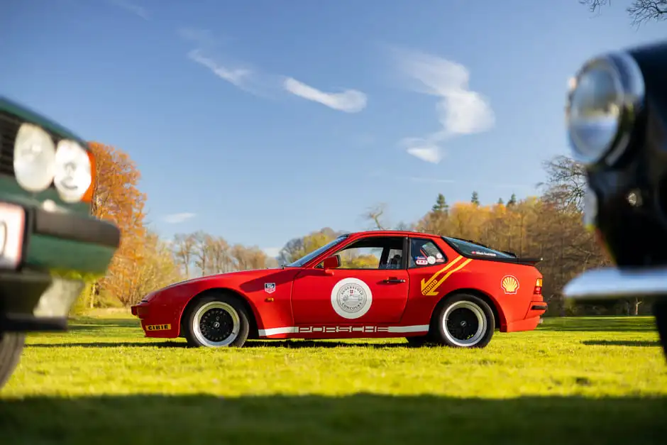 A red Porsche 944 on the estate lawn, framed between two other classic cars, under a clear autumn sky.