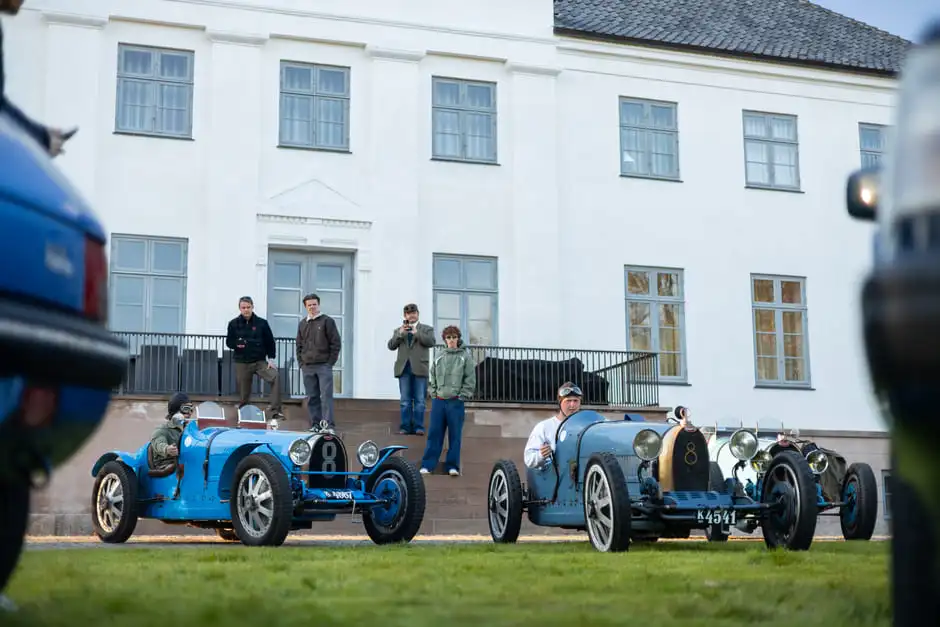 Two blue Bugatti racers before the Aggersvold manor, drivers seated and onlookers on the steps behind.